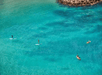 Four people are paddleboarding and kayaking in clear, turquoise water near a rocky shoreline.