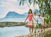 Four children walk in a line on a stone path by the beach, with palm trees and mountains in the background.