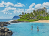 Two people paddleboarding on clear blue water near a beach with palm trees and a hotel in the background. Cloudy sky and distant mountain visible.