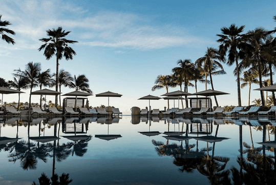A serene pool scene with palm trees and lounge chairs reflecting in the water under a clear sky.
