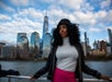 A person in a black jacket and pink skirt stands on a boat with the New York City skyline and One World Trade Center in the background.