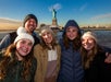 A group of five people wearing winter clothing smile in front of the Statue of Liberty on a clear day.
