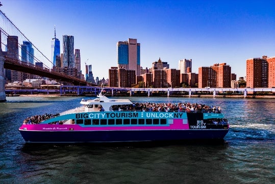A teal and pink cruise boat with the top deck stuffed with people with the city in the background and the Brooklyn bridge to the left.