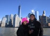 A couple stands in front of the New York City skyline, featuring the One World Trade Center. The woman wears a pink hat; both are dressed in black. The sky is clear and sunny.