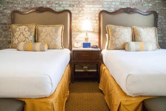 Two neatly made beds with decorative pillows and bolsters, separated by a nightstand with a lamp. The wall behind is brick-patterned, and the beds have wooden headboards.