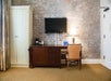 Room interior with a brick wall, a mounted flat-screen TV, wooden desk, chair, table lamp, coffee maker, and cabinet.