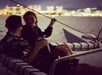 Two people sit on a boat at night, facing each other and talking. City lights and skyline are visible in the blurred background across the water.