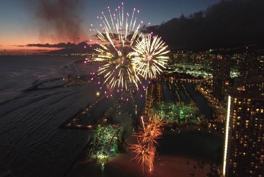 Fireworks light up the night sky above a coastal city, with ocean waves and illuminated buildings visible below.
