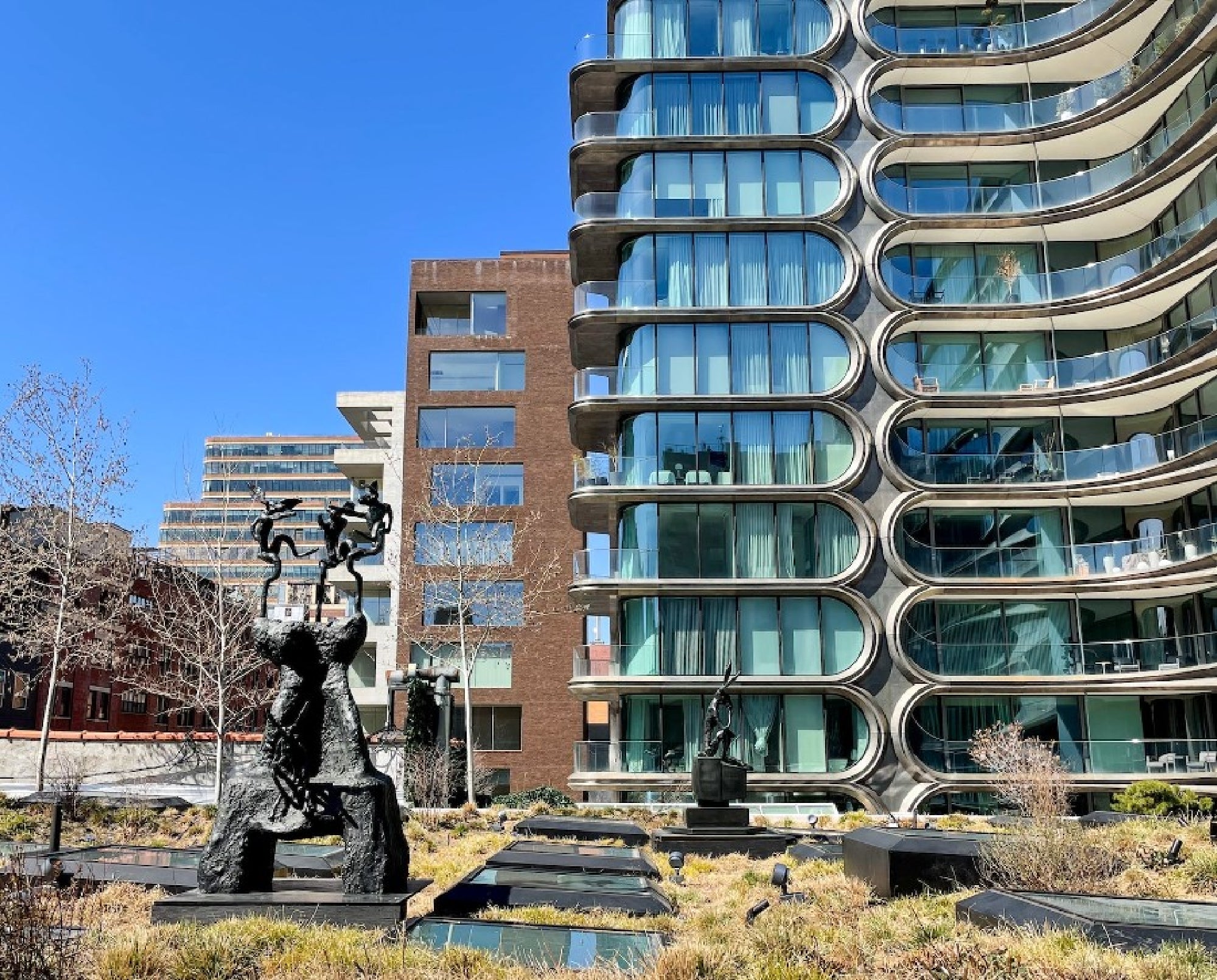 Modern residential buildings with curved balconies stand next to abstract sculptures in a landscaped area under a clear blue sky.