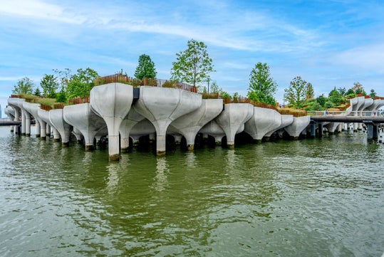 Elevated concrete structure resembling flower petals, topped with trees and greenery, extends over a body of water under a blue sky.
