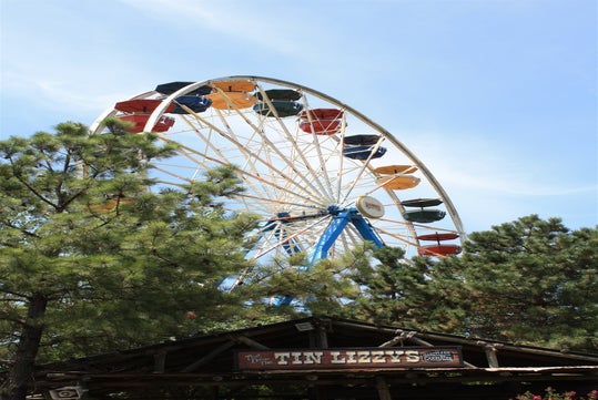 A Ferris wheel with colorful gondolas rises above green trees, with a rustic wooden sign reading "Tin Lizzy's" in the foreground.