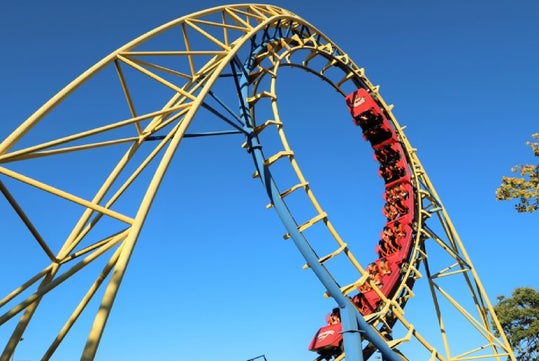 A roller coaster with a yellow and blue loop, carrying passengers through an upside-down loop against a clear blue sky.