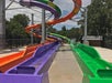 Colorful water slides winding down a structure, featuring purple, orange, and green slides, surrounded by trees and a metal fence.