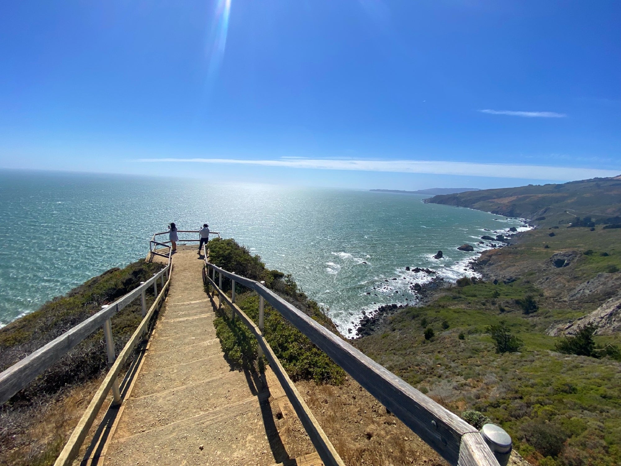 A paved pathway with wooden railings leads to a scenic overlook above the ocean, with two people standing at the end, facing the coastline and water.