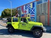 Two women pose in a bright green Jeep parked on a street, with one standing through the open roof. Behind them is a wall decorated with colorful nautical flag designs.