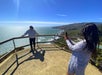One person takes a photo of another person standing by a wooden railing on a coastal cliff overlooking the ocean under a clear blue sky.