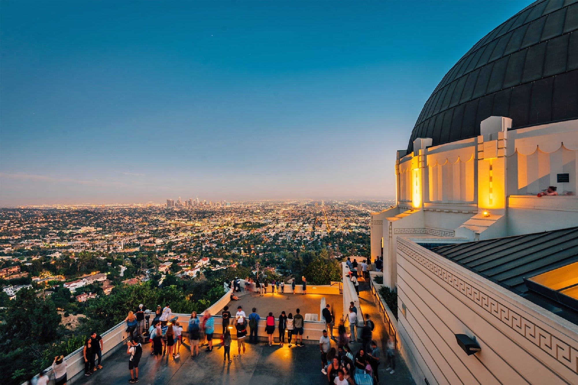 People stand on an observation deck at Griffith Observatory at dusk, overlooking the city lights of Los Angeles with downtown visible in the distance.