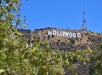 The Hollywood sign on a dry hillside under a clear blue sky, with green foliage in the foreground and a tall radio tower nearby.