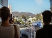 Two people view and photograph the Hollywood sign from a balcony overlooking a busy street with billboards and palm trees in Los Angeles.