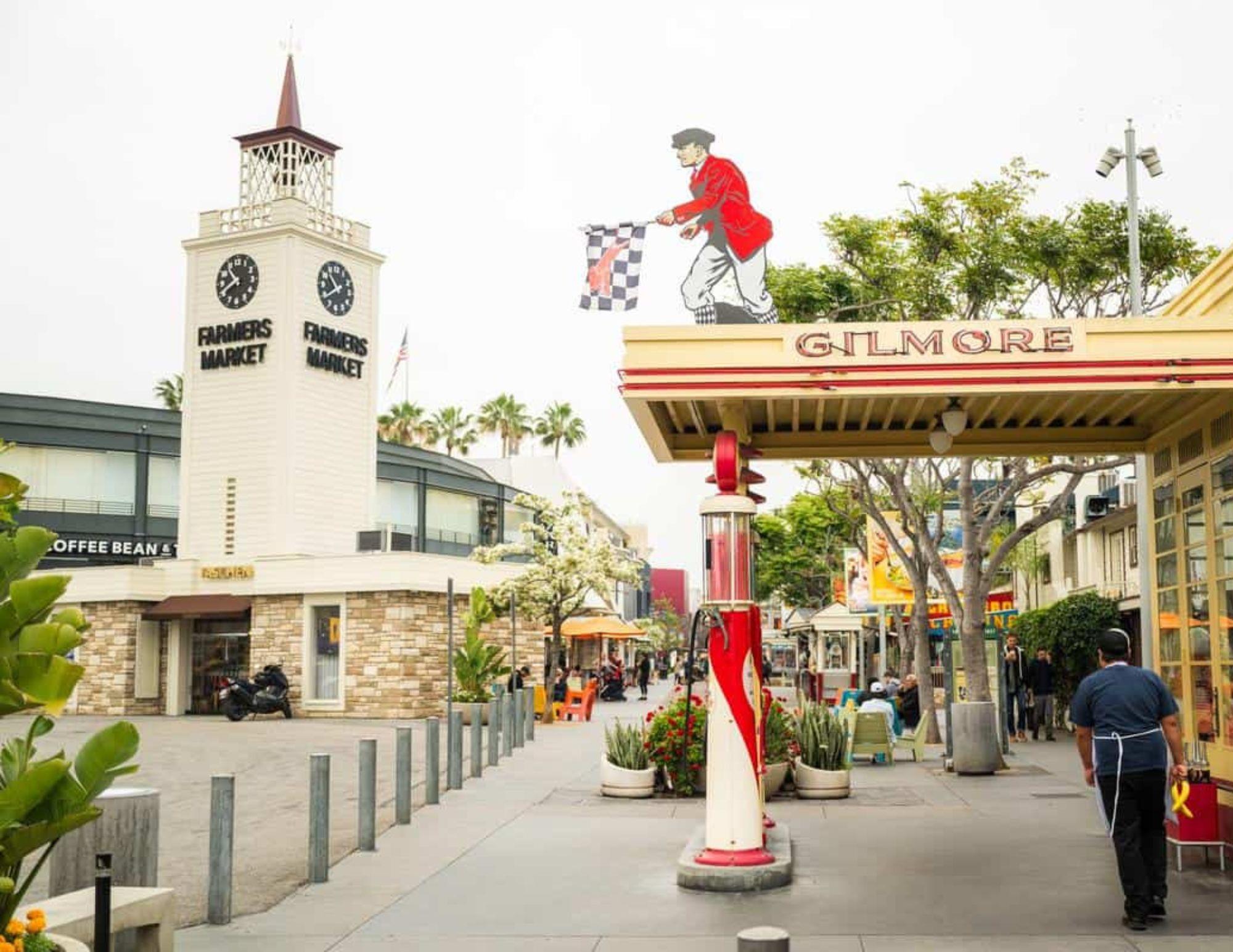 Outdoor scene at the Los Angeles Farmers Market, showing a clock tower, Gilmore gas pump, and people walking near shops and cafes.
