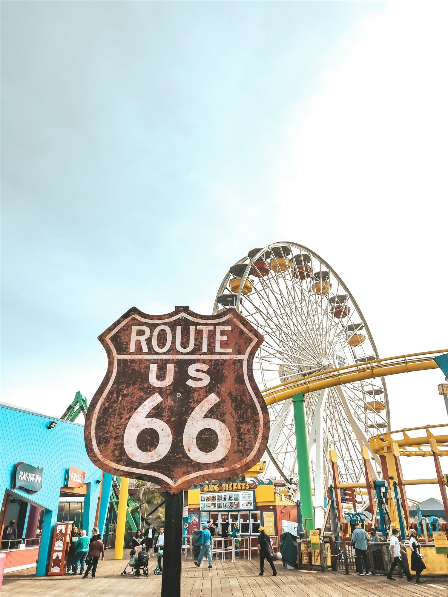 A Route 66 sign stands in front of a Ferris wheel and colorful amusement park buildings on a boardwalk with several people walking nearby.