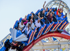 People of various ages ride a blue and red roller coaster on a sunny day, some smiling and raising their hands as the ride descends a curved track.