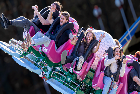 Four people ride a roller coaster with pink seats, smiling and raising their arms as the ride tilts to one side, with blurred background and a large decorative sphere behind them.