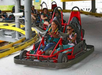 Two people drive a red go-kart on a track, both wearing seat belts and smiling. Other riders in go-karts are visible in the background.