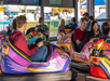 People riding colorful bumper cars indoors at an amusement park, interacting and smiling, with other park attractions visible in the background.
