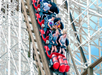 A group of people ride a red roller coaster down a steep wooden track, with some passengers' hair blown upwards by the speed.
