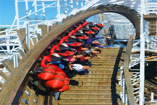 A roller coaster train with riders tilts sideways on a wooden track, with passengers raising their arms and smiling as they go through a steep curve.
