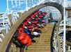 A roller coaster train with riders tilts sideways on a wooden track, with passengers raising their arms and smiling as they go through a steep curve.