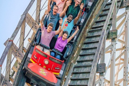 A group of people ride a wooden roller coaster, holding their arms up as the car descends a steep track.