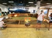 Three people observe reptiles in wooden enclosures at the Idaho Reptile Zoo, with a large alligator mural and informative sign visible in the background.