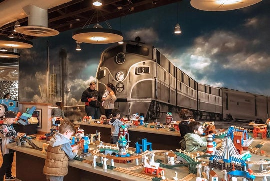Children play with toy train sets on tables in a room with a large mural of a vintage locomotive on the wall. Several adults stand nearby. The space is well-lit and busy.