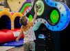 A young boy reaches up to play an arcade game with colorful lights and digital displays in an indoor play area.