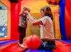 A woman helps a toddler balance while standing on a colorful inflatable bounce house with a red basketball in front of them.