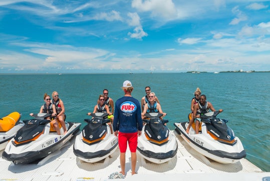 A group of people on jet skis listens to an instructor in a "Fury" shirt on a sunny day by the ocean.