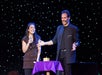 A magician performs on stage next to a smiling woman holding a microphone. The background is decorated with small lights.