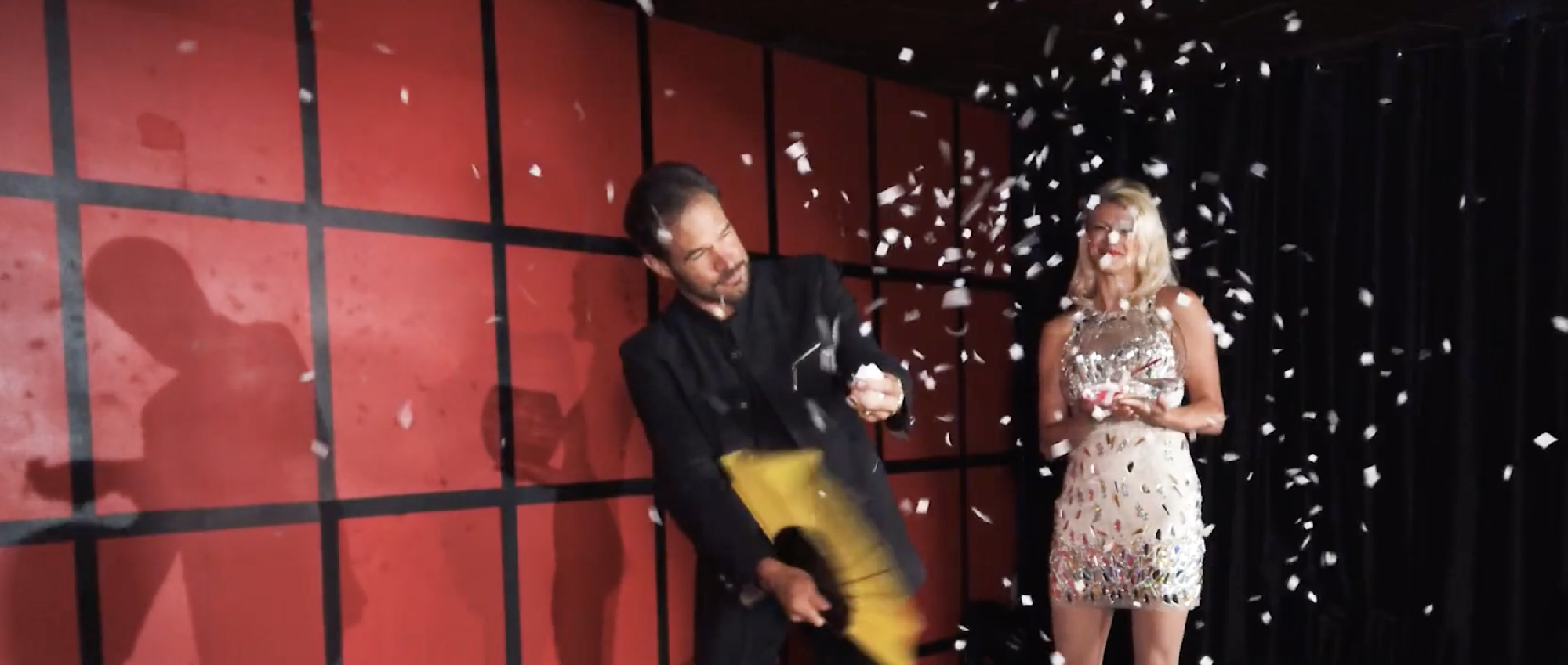Magician performs with a yellow umbrella, creating a flurry of paper scraps, as a woman in a sparkly dress watches on a red and black stage background.