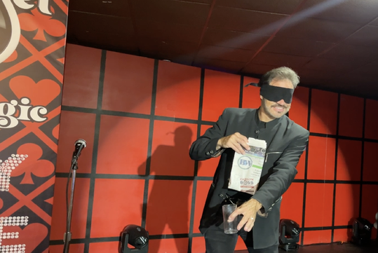 A blindfolded man in black pours liquid from a carton into a glass on stage. The backdrop features red hearts and patterns.