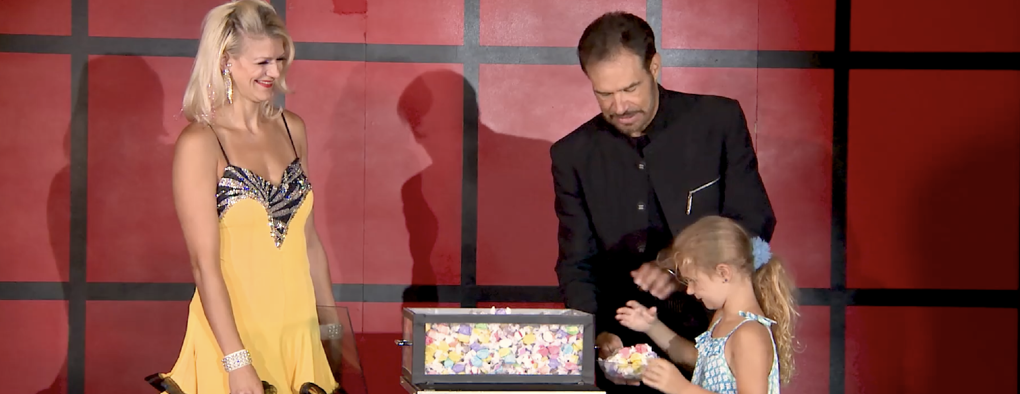 A man in a black suit stands beside a woman in a yellow dress and a child. The child looks at colorful pieces in a clear box on a table. A red and black wall is in the background.