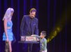 A magician and an assistant perform a trick on stage with a young boy selecting an item from a box, under stage lights and a blue backdrop.