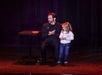 A man kneels on stage next to a young girl. They are facing an audience, with a small table beside them. The man is clapping and wearing a headset microphone.