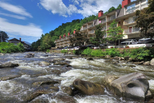 A scenic view of a fast-flowing river with large rocks in the foreground, flanked by lush green trees and a multi-story building with red chimneys and balconies on the right side, under a bright blue sky with scattered clouds.