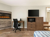 A guest room corner with a desk, office chair, coffee maker, electric fireplace, TV on a dresser, microwave, and partial view of a bed.