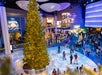 A large decorated Christmas tree stands in the center of a crowded indoor venue with festive lights and people walking around.