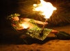 A person in traditional Polynesian attire performs a fire dance at night, holding a flaming torch and surrounded by palm leaves on a sandy surface.