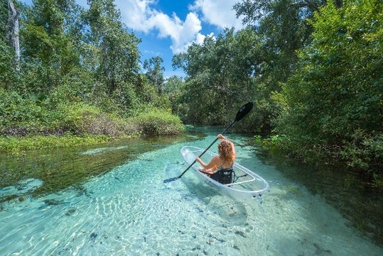 A person paddles a clear kayak upstream through Rock Springs Run under a blue sky.