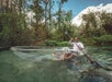 A man paddles a clear kayak over spring water surrounded by greenery.